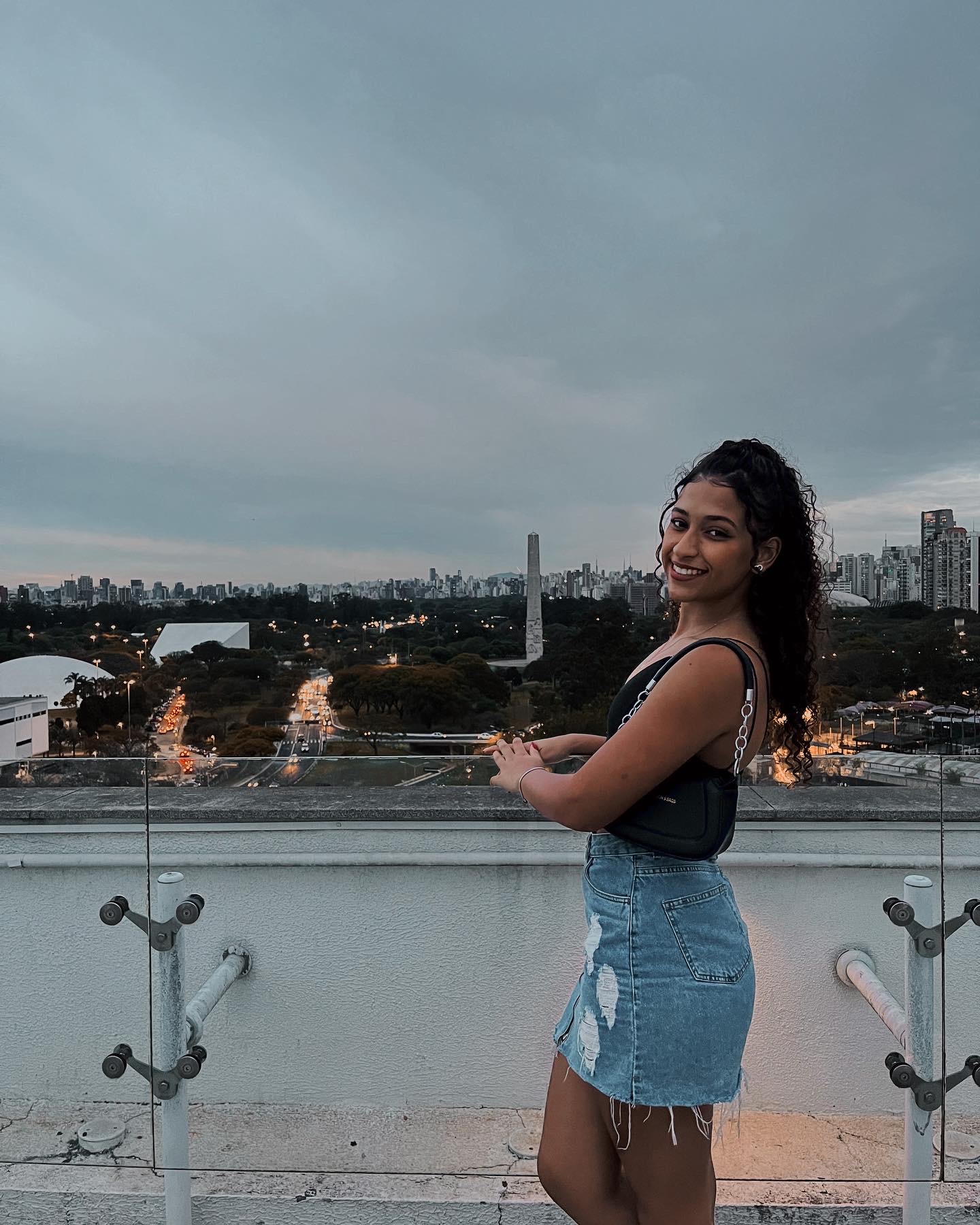 Foto de Carol no topo de um edifício, ela está de saia jeans, top e bolsa pretos. Sorri para câmera e se apoia em muro de vidro